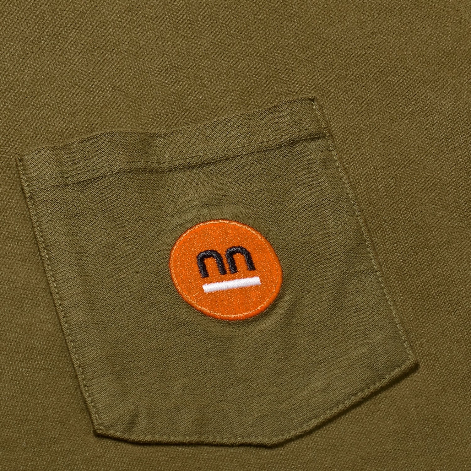 Close-up of the khaki Berlin Tee's pocket with orange Pamuuc logo.